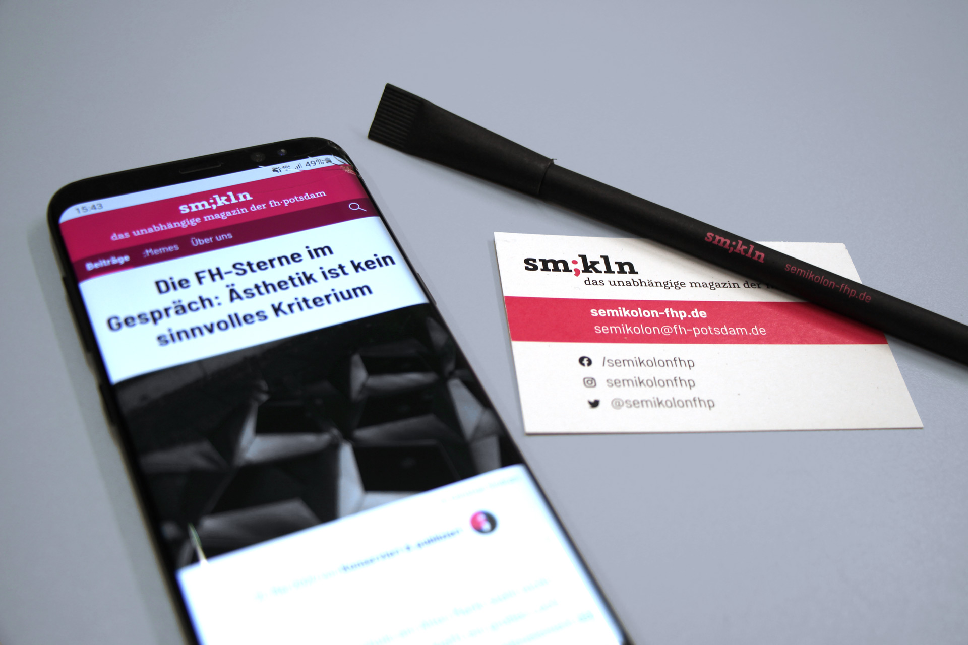 Eine Visitenkarte und ein Kugelschreiber im Design von Semikolon, daneben zeigt ein Smartphone die Semikolon-Webseite